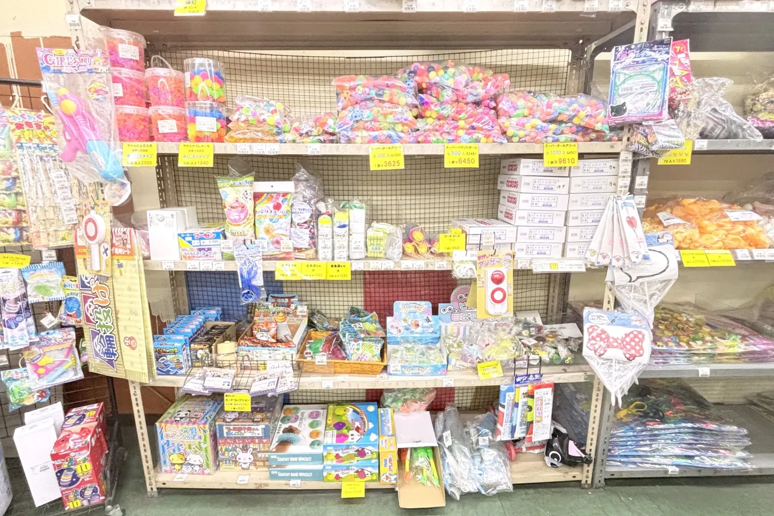 スーパーボール・水ヨーヨー・水鉄砲・金魚すくいの袋・景品のおもちゃが、値札とともに棚いっぱいに並ぶ店内の縁日・景品用品の売り場